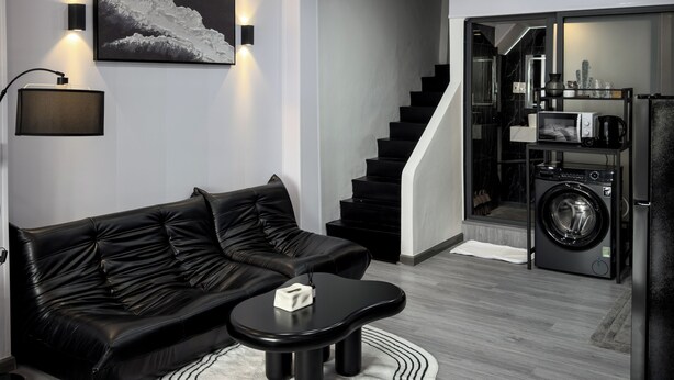 Een moderne woonkamer met een zwarte leren bank, een ronde salontafel en een zwart tapijt. Een trap leidt naar een keuken met een wasmachine en een magnetron.