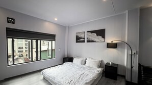 Een moderne slaapkamer met een groot bed, een raam met uitzicht op gebouwen, een vloerlamp en twee kunstwerken aan de muur.