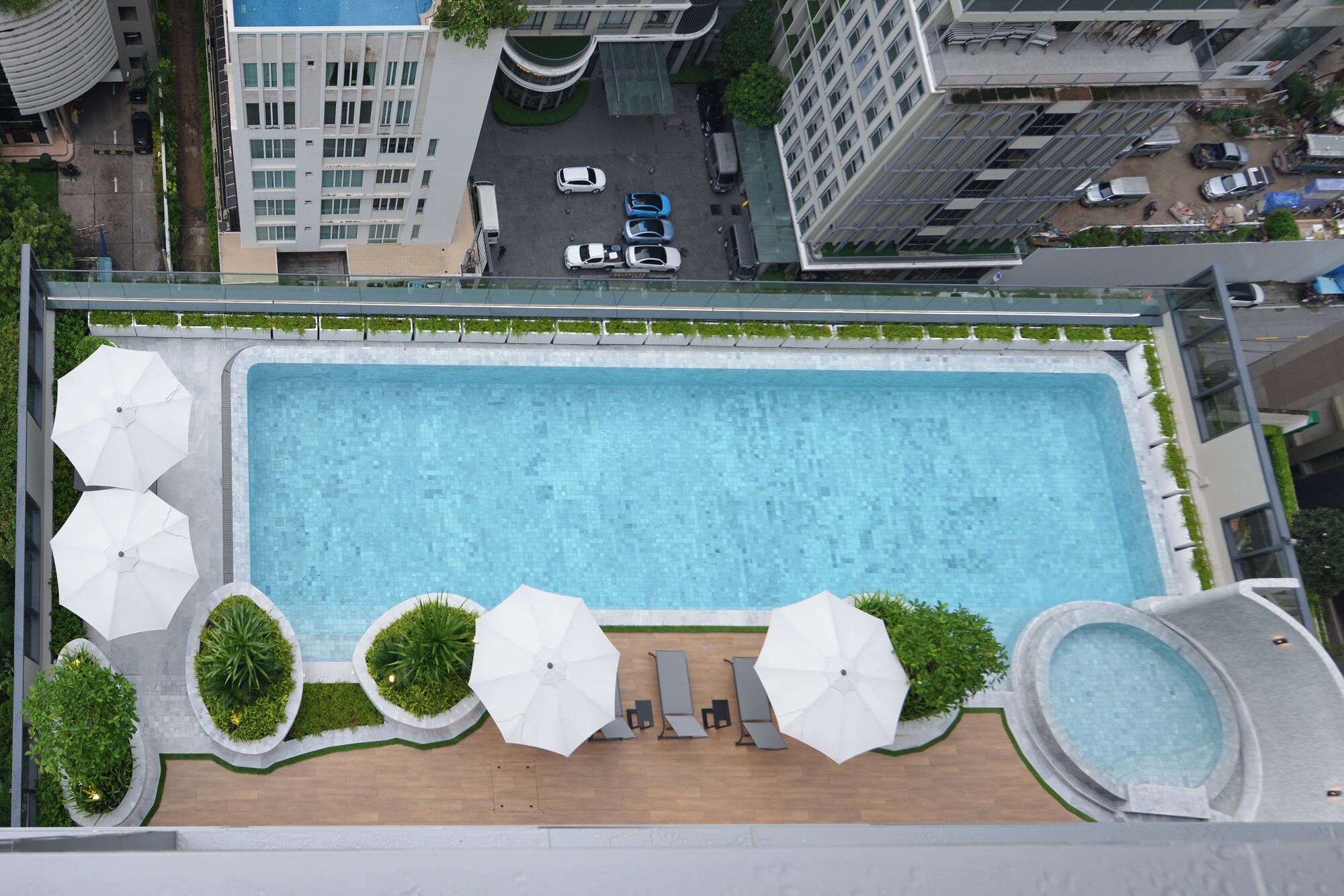 An aerial view of a rooftop swimming pool with multiple umbrellas, surrounded by greenery and adjacent to high-rise buildings.