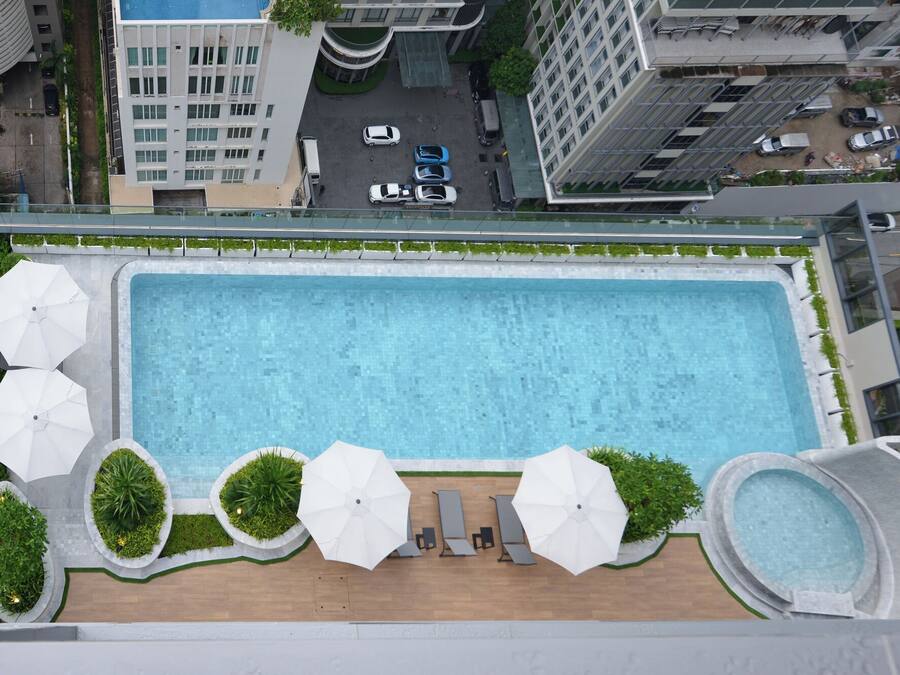 An aerial view of a rooftop swimming pool with multiple umbrellas, surrounded by greenery and adjacent to high-rise buildings.