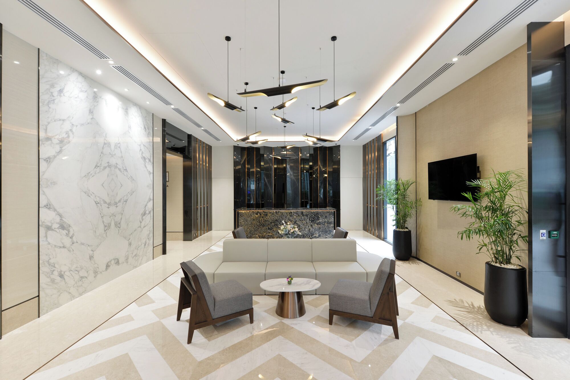 A modern lobby with a marble reception desk, a sofa, two armchairs, a round coffee table, a large potted plant, a flat-screen TV, and an elevator.