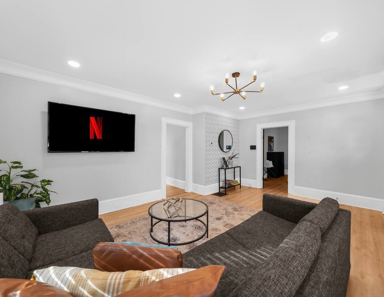 A modern living room with a flat-screen TV, grey sofas, a glass coffee table, and a chandelier.