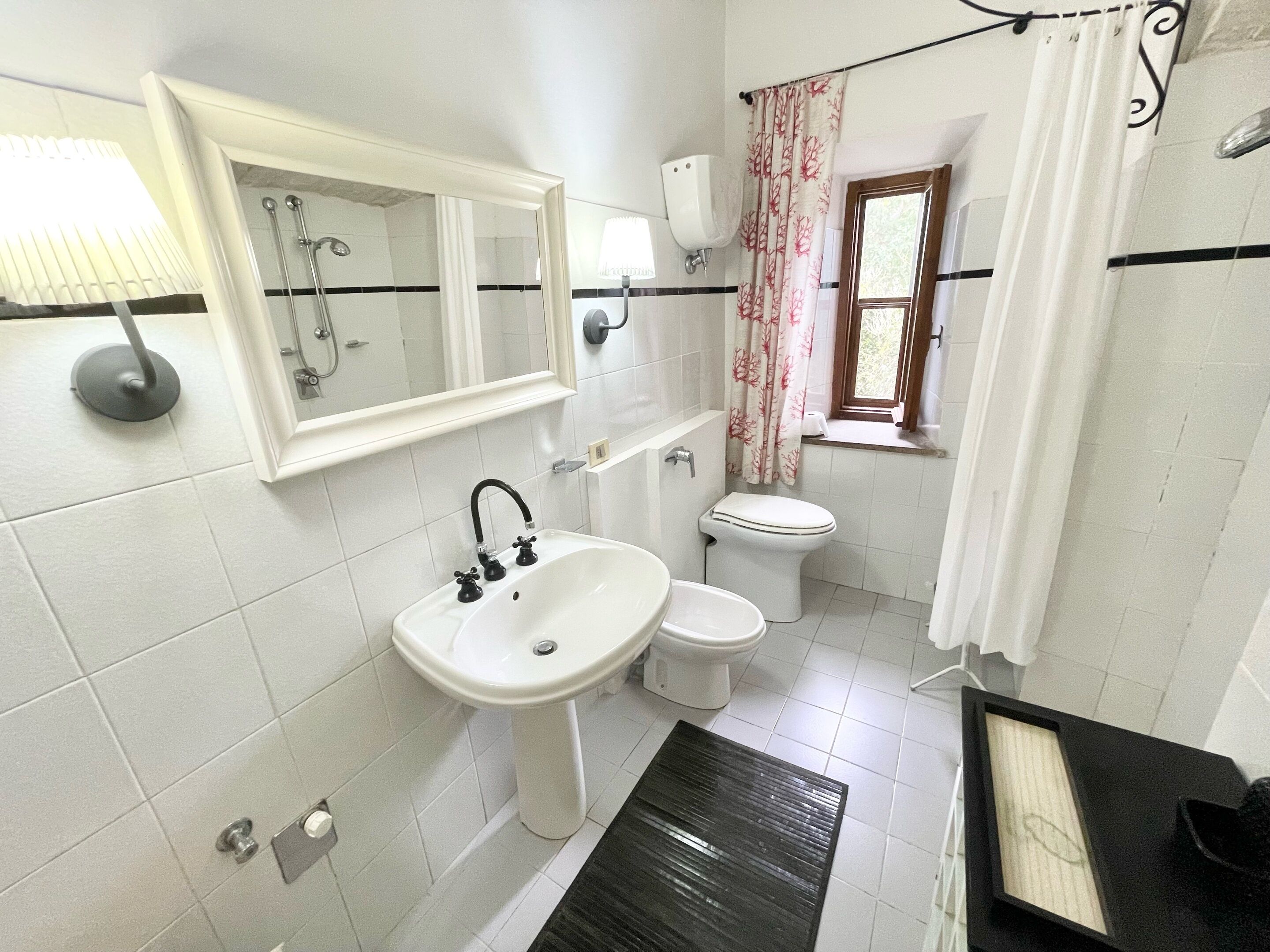 Un baño con lavamanos blanco, inodoro, ducha y ventana con cortina.