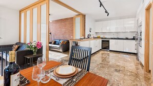 A modern kitchen with white cabinets, a dining area with a wooden table, and a living room with a sofa and a brick wall. - Solaga - Mary (Málaga)
