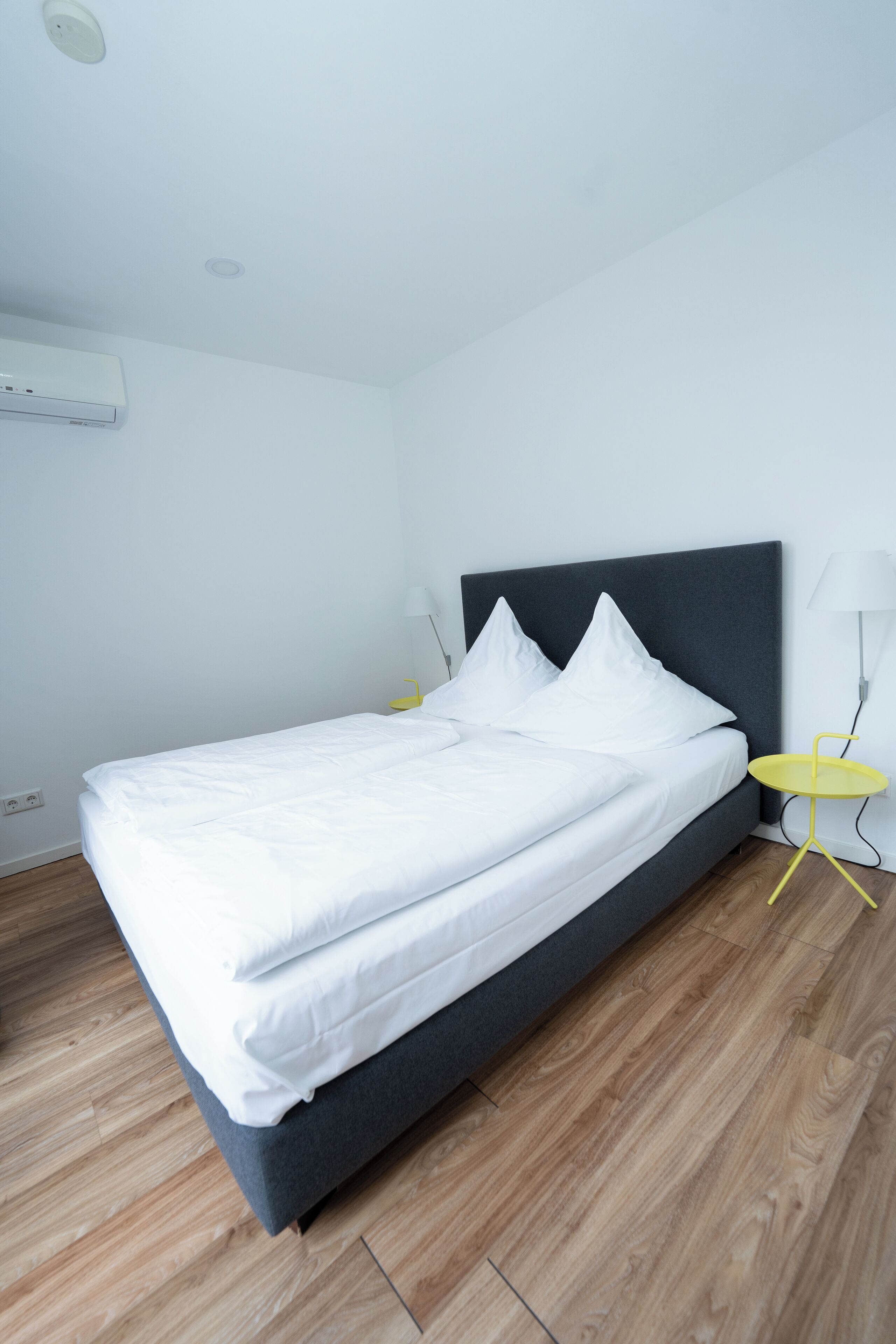 A neatly made bed with white linens, a grey headboard, and a yellow bedside table with a lamp.