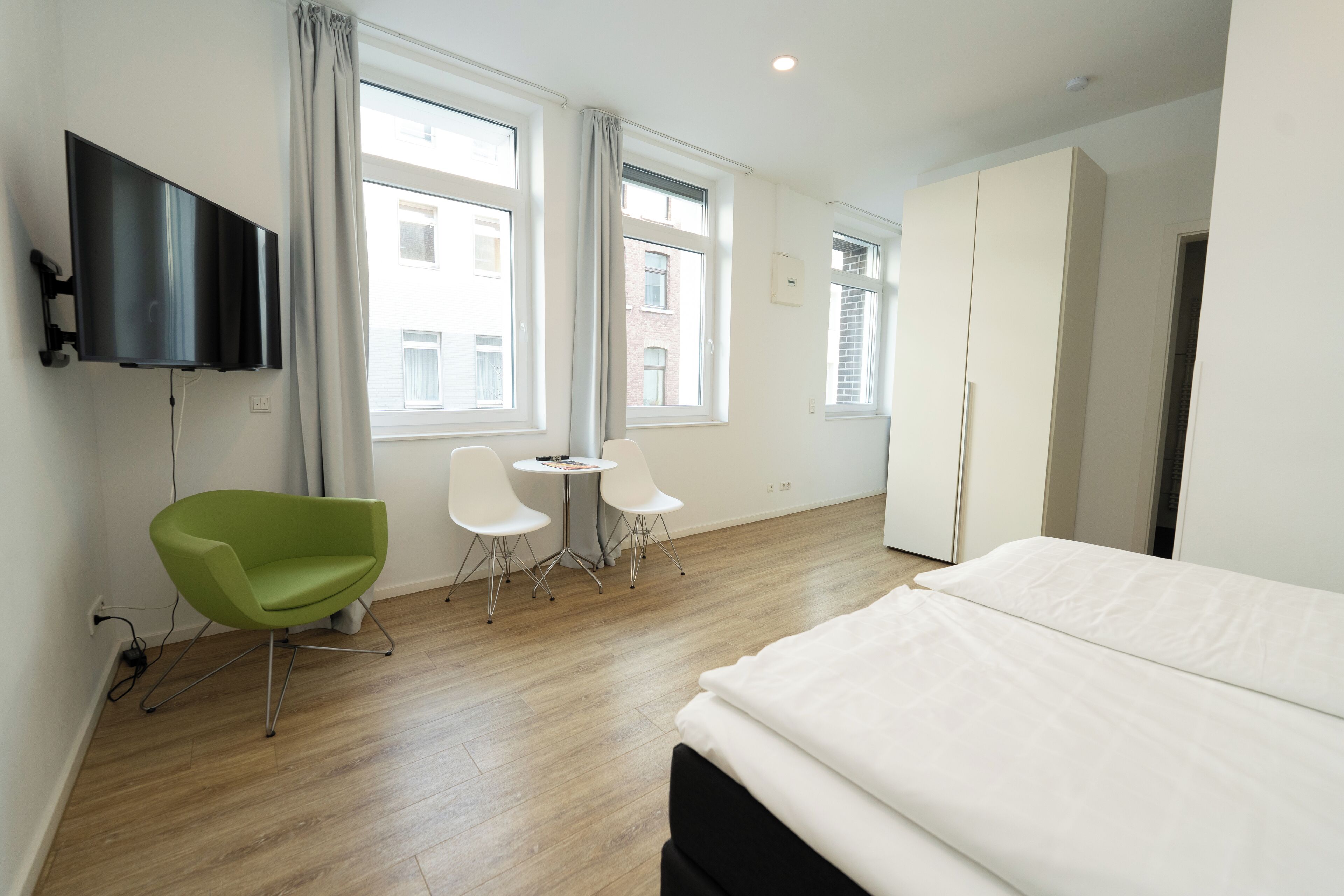 Een hotelkamer met een bed, een aan de muur bevestigde televisie, twee stoelen, een klein tafeltje en grote ramen.