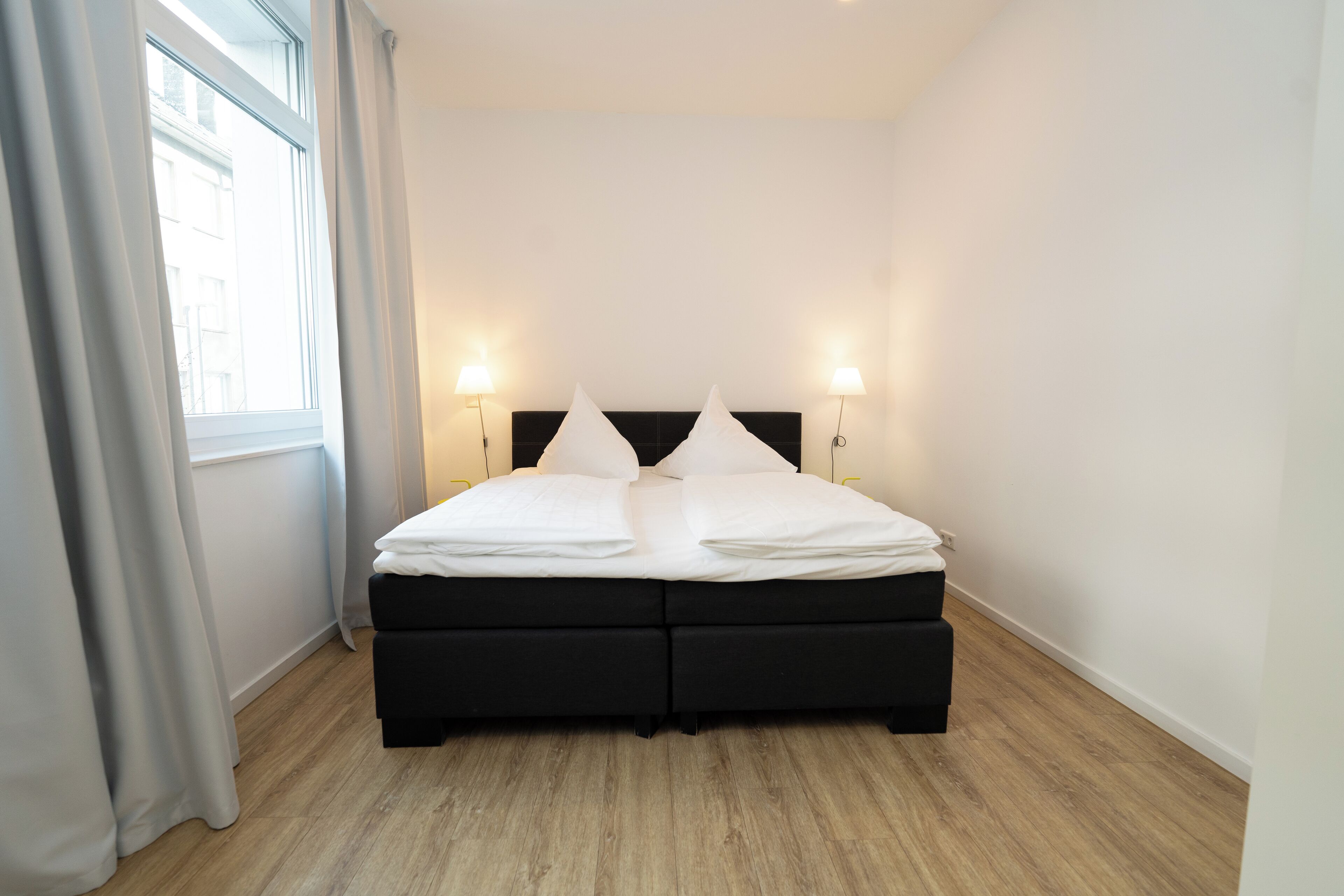 A modern hotel room with a black bed, white linens, two bedside lamps, and a view through a window with curtains.