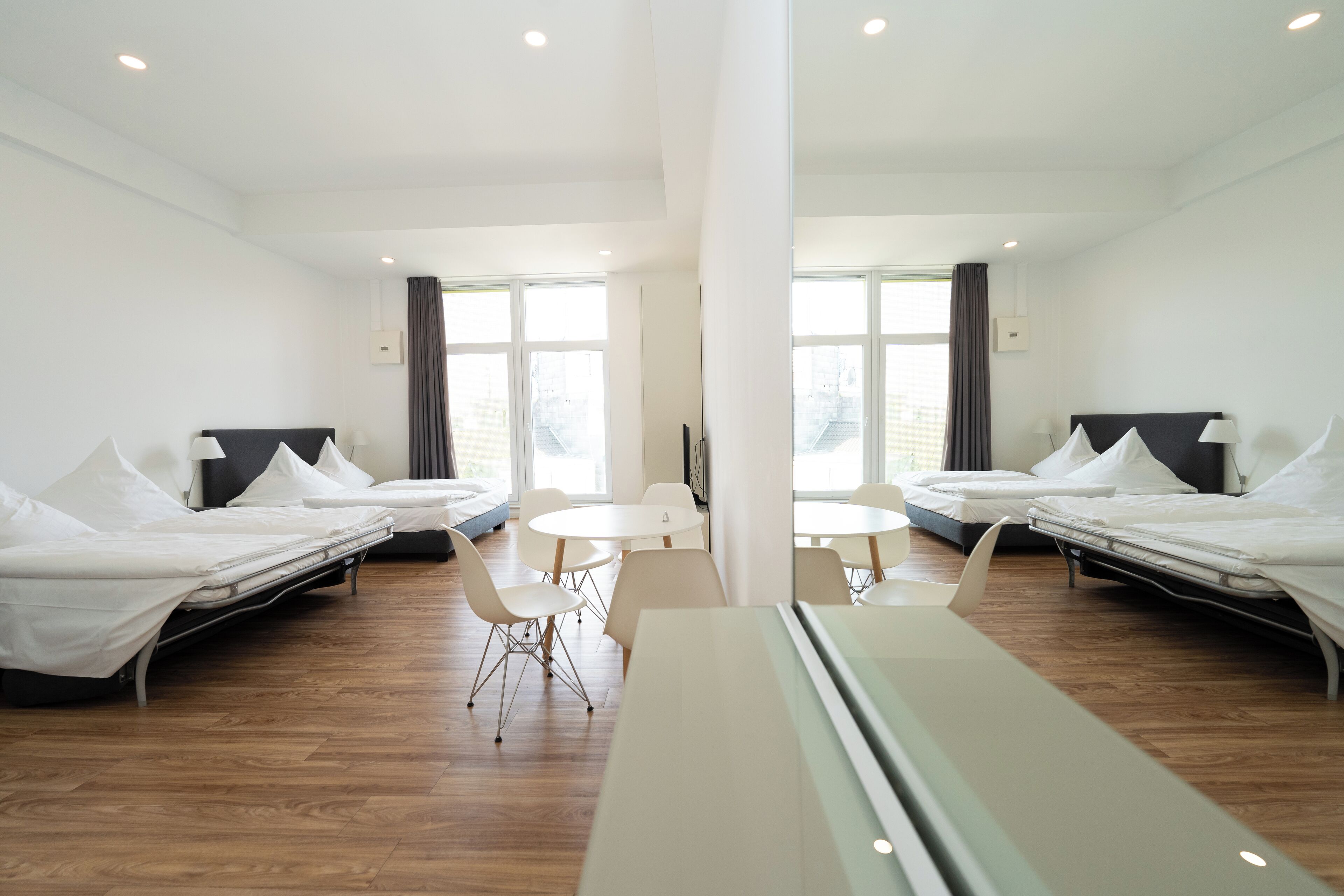 A modern hotel room with two single beds, a small dining table, and a large window providing natural light.