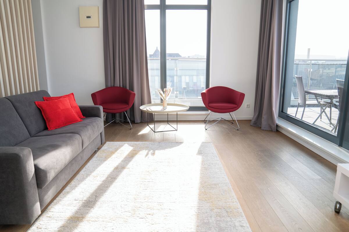 A modern living room with a grey sofa, red chairs, a small round table, and a large window offering a city view.