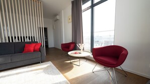 A modern living room with a grey sofa, red chairs, a round coffee table, and a large window with a view of the city. - Das Loft Apartments (Aachen)