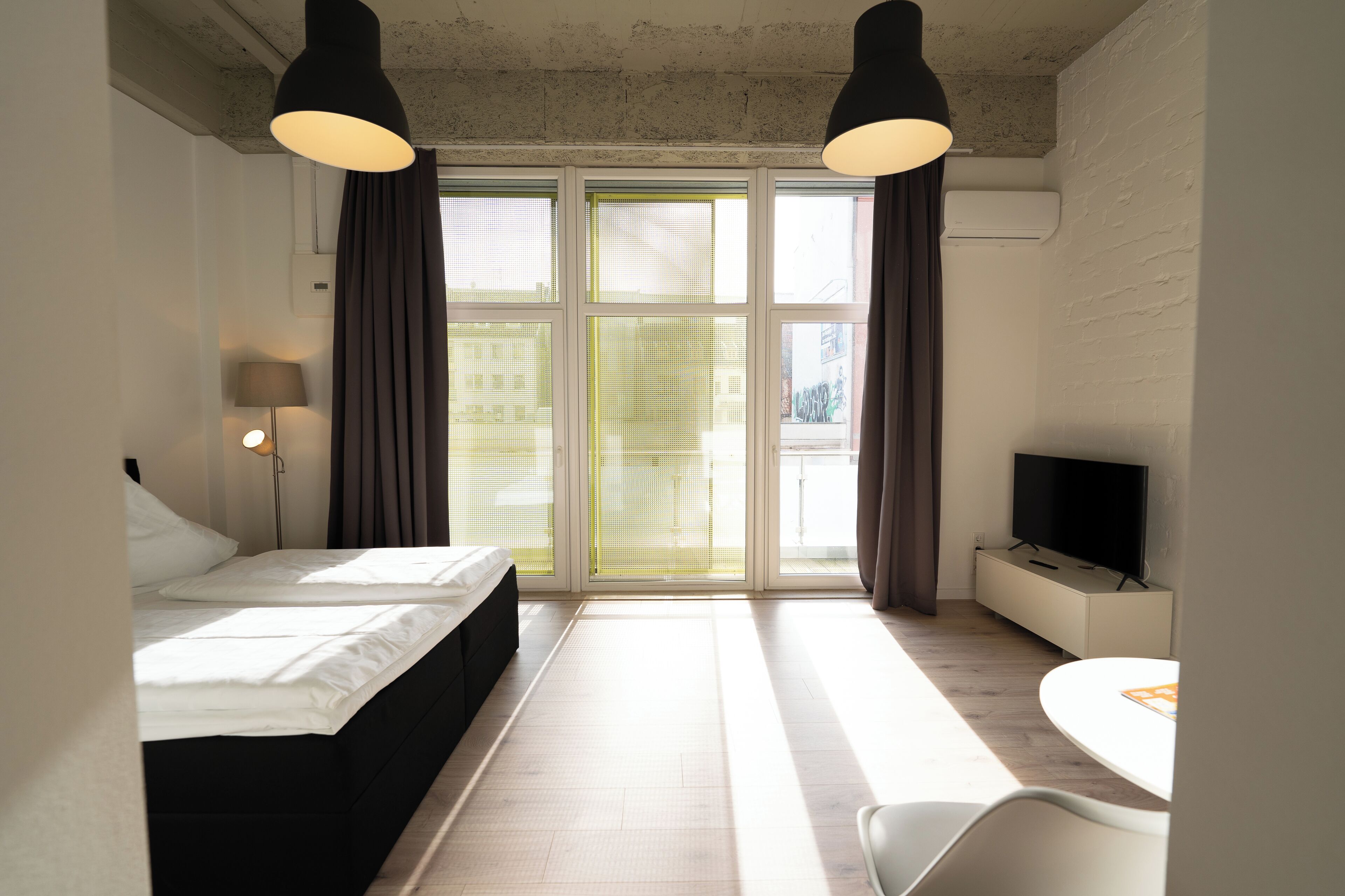A modern hotel room with a bed, a desk, a TV, and large windows with curtains.