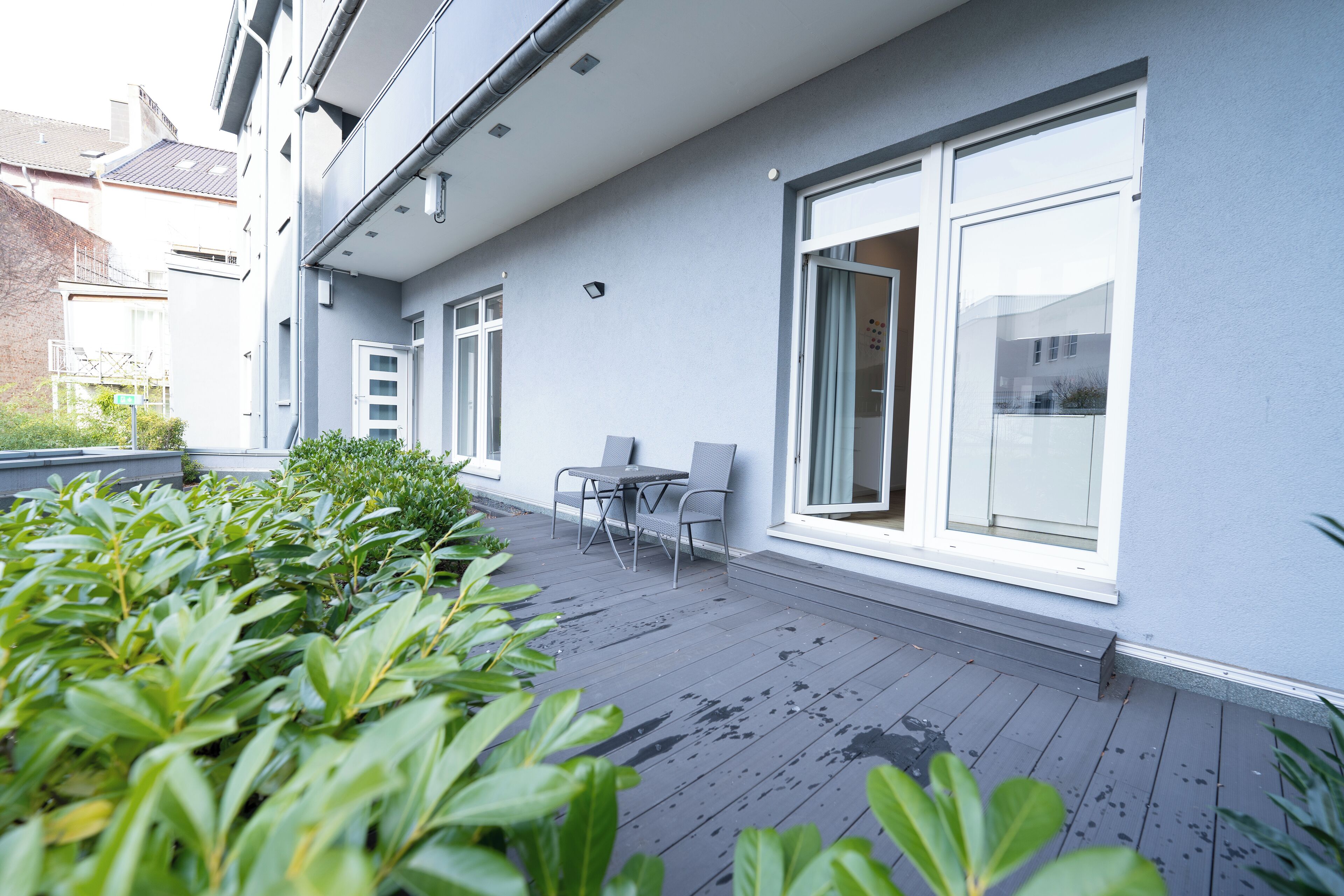 Un patio moderno al aire libre con cubierta de madera gris, una zona de estar con mesa y sillas, y ventanas amplias con vistas a los edificios circundantes.