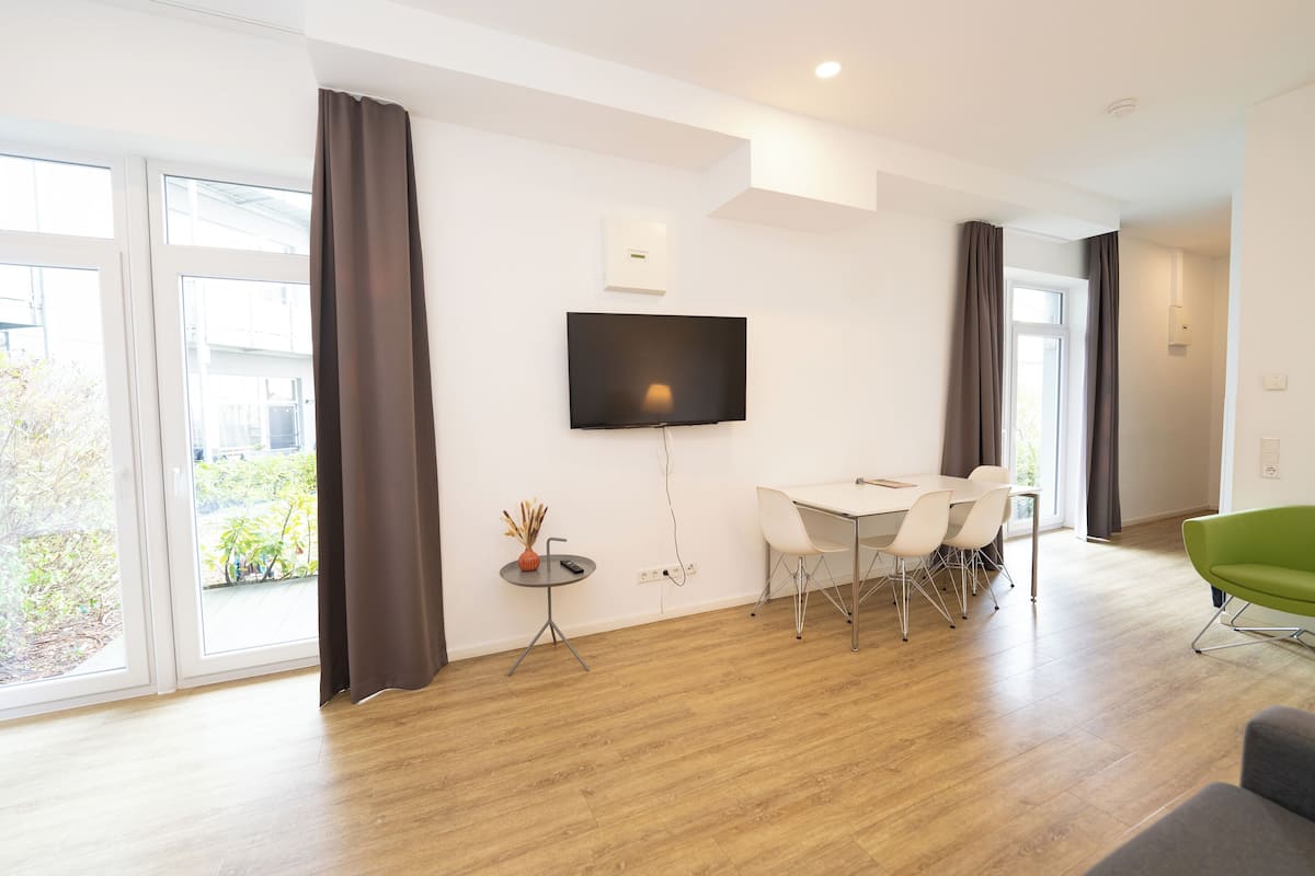 A modern living room with a flat-screen TV mounted on the wall, a dining table with chairs, and a green armchair.