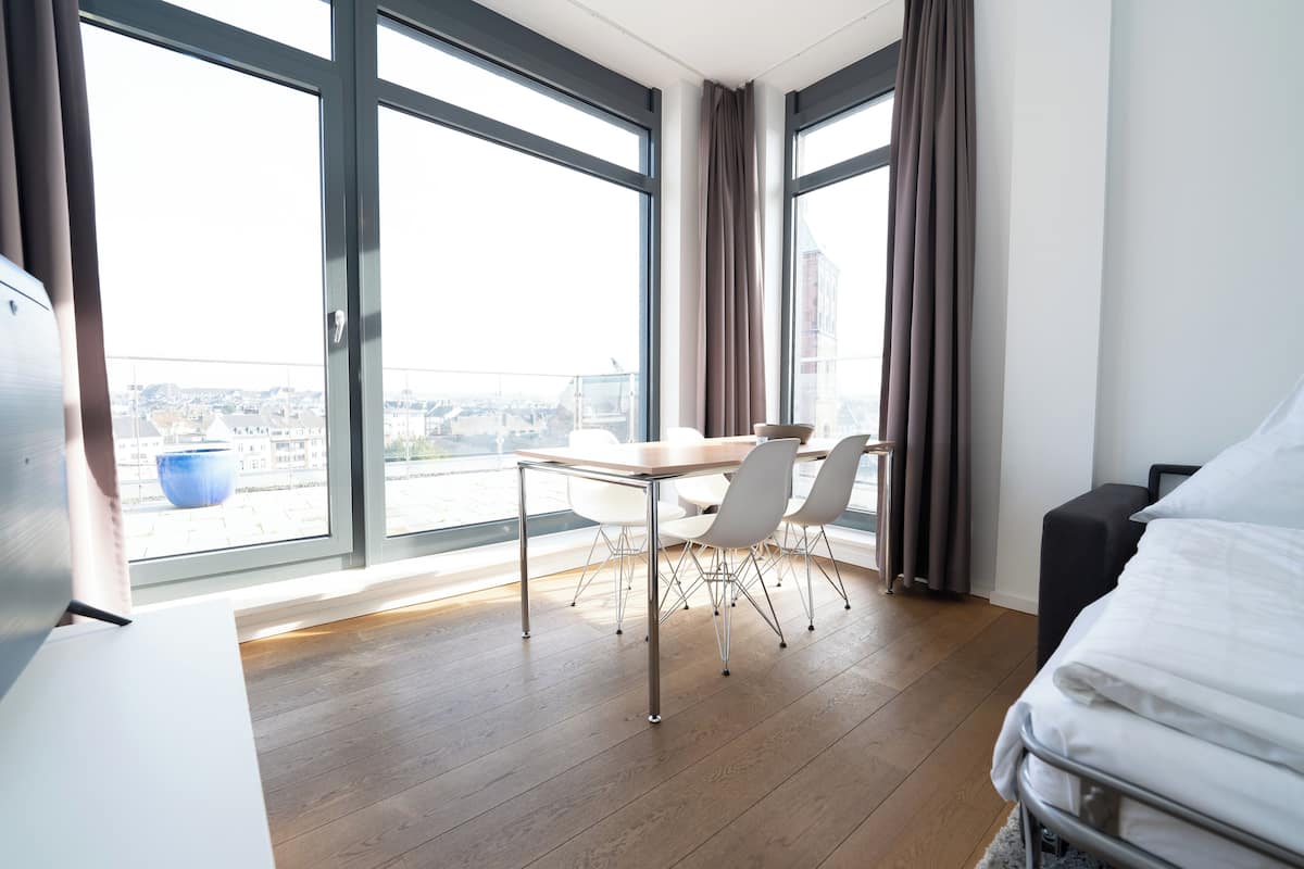 A modern hotel room with a large window, a dining table with chairs, a bed, and a small black ottoman.