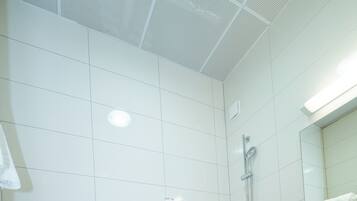 Un baño con paredes de azulejos blancos, suelo de losetas negras, inodoro blanco, ducha con cabezal de ducha manual y lavamanos blanco con espejo encima.