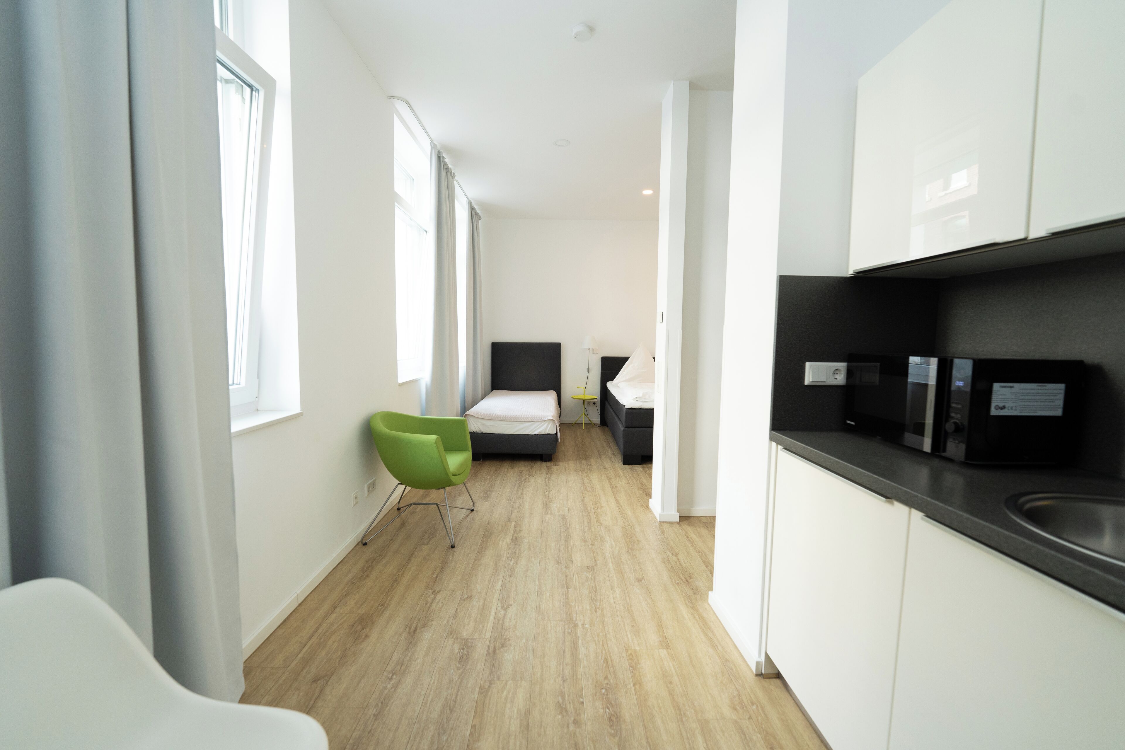 Una habitación de hotel moderna con una cama, una silla verde, una mesita y una kitchenette con fregadero y microondas.