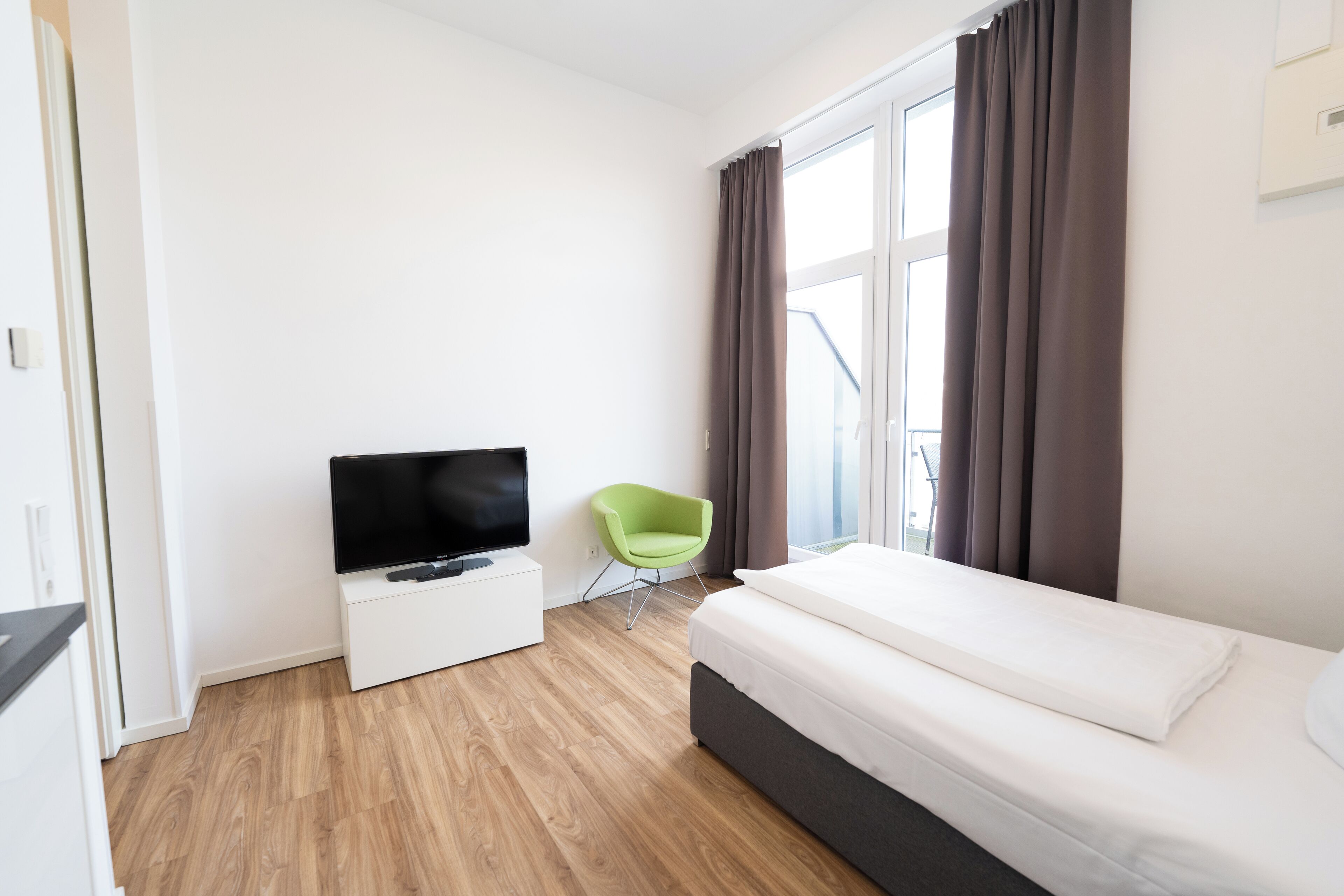 A hotel room with a bed, a TV on a white cabinet, a green chair, and a window with curtains.
