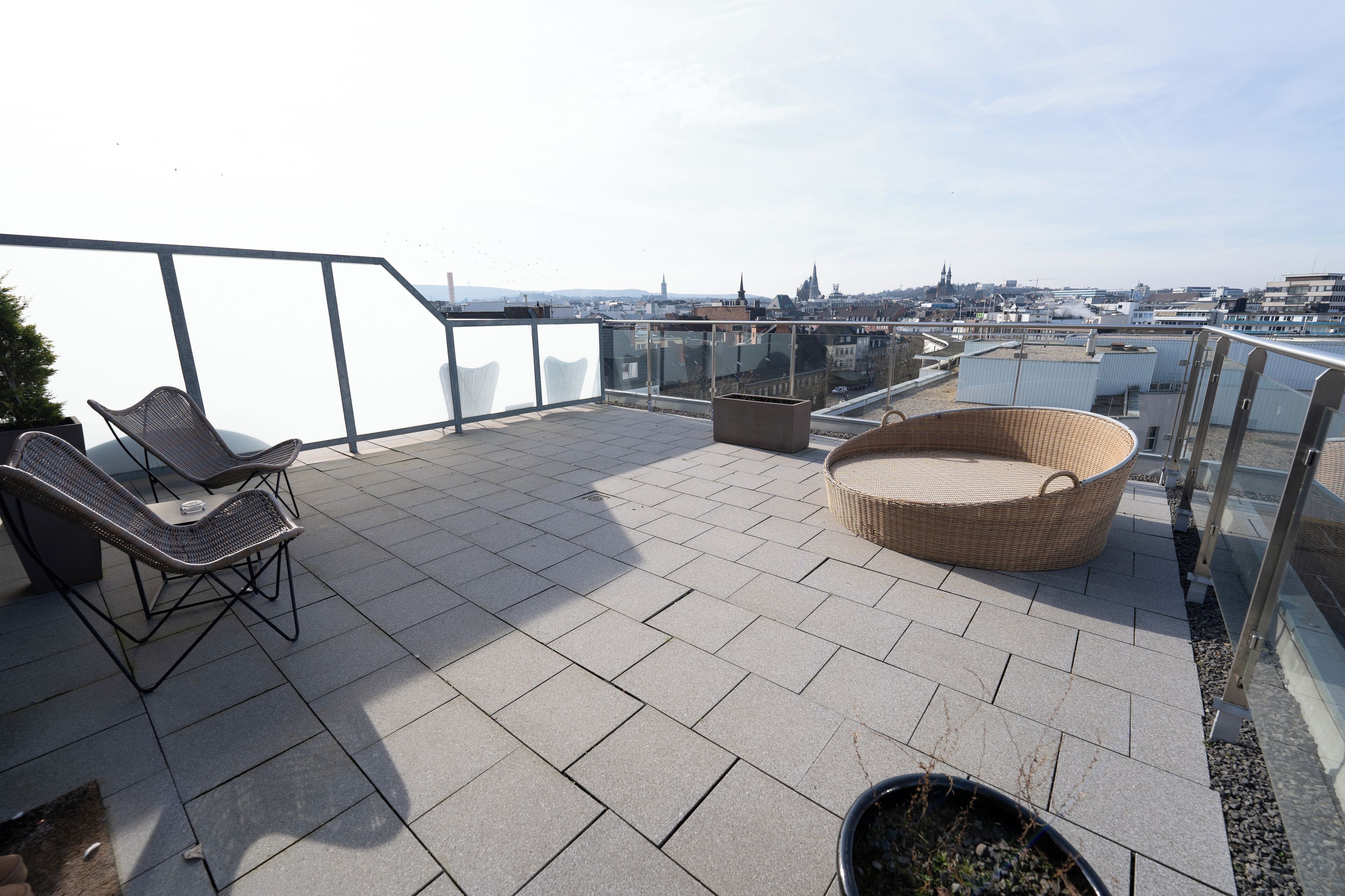 A rooftop terrace with a view of the city, featuring a wicker chair and a cushioned seating area.
