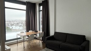 A modern living room with a sofa, a dining table with chairs, and a large window offering a city view. - Das Loft Apartments (Aachen)