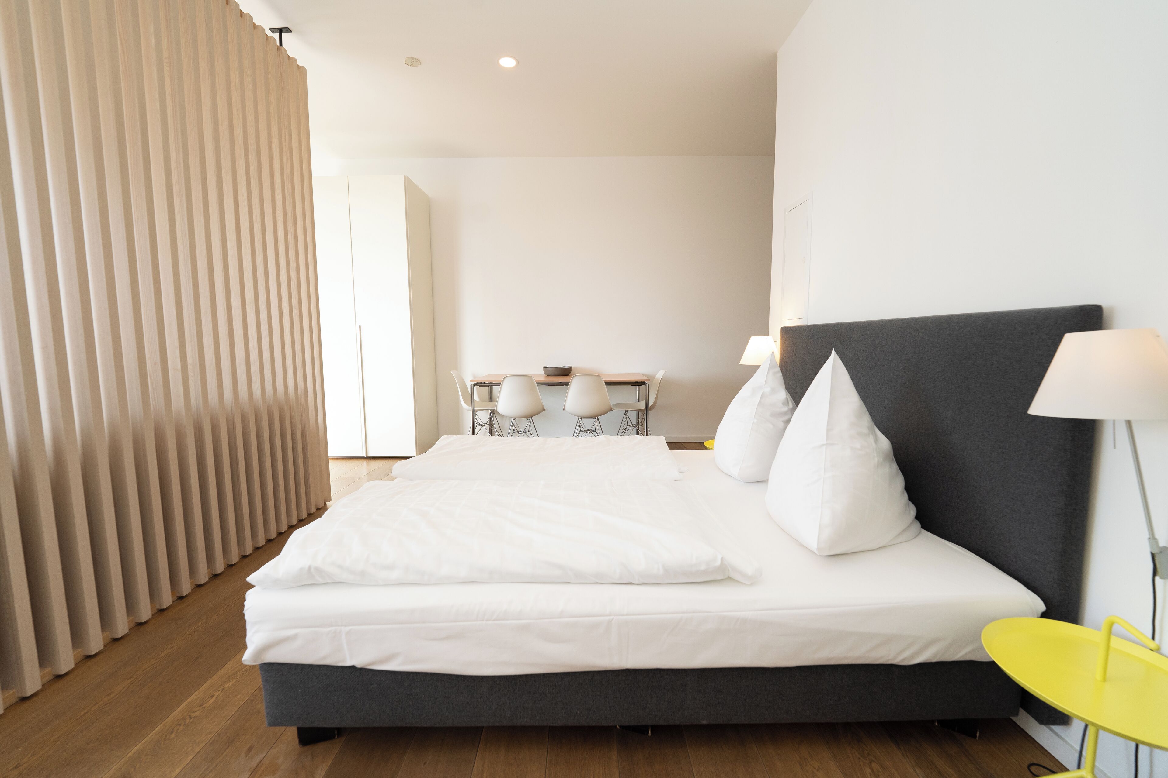 A modern hotel room with a large bed, a bedside table, a lamp, and a view of a dining area.
