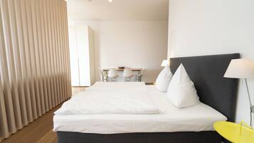 A modern hotel room with a large bed, a bedside table, a lamp, and a view of a dining area.