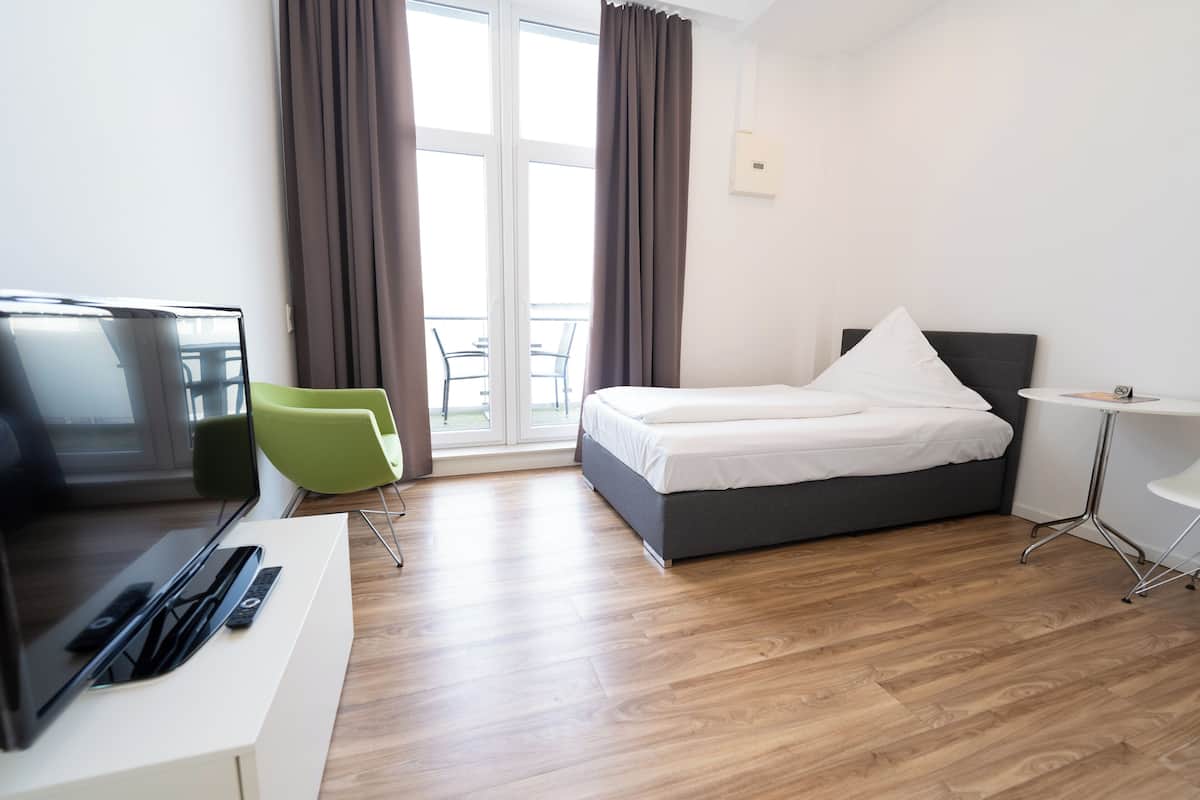 A modern hotel room with a bed, a desk with a flat-screen TV, a green chair, and a small table.