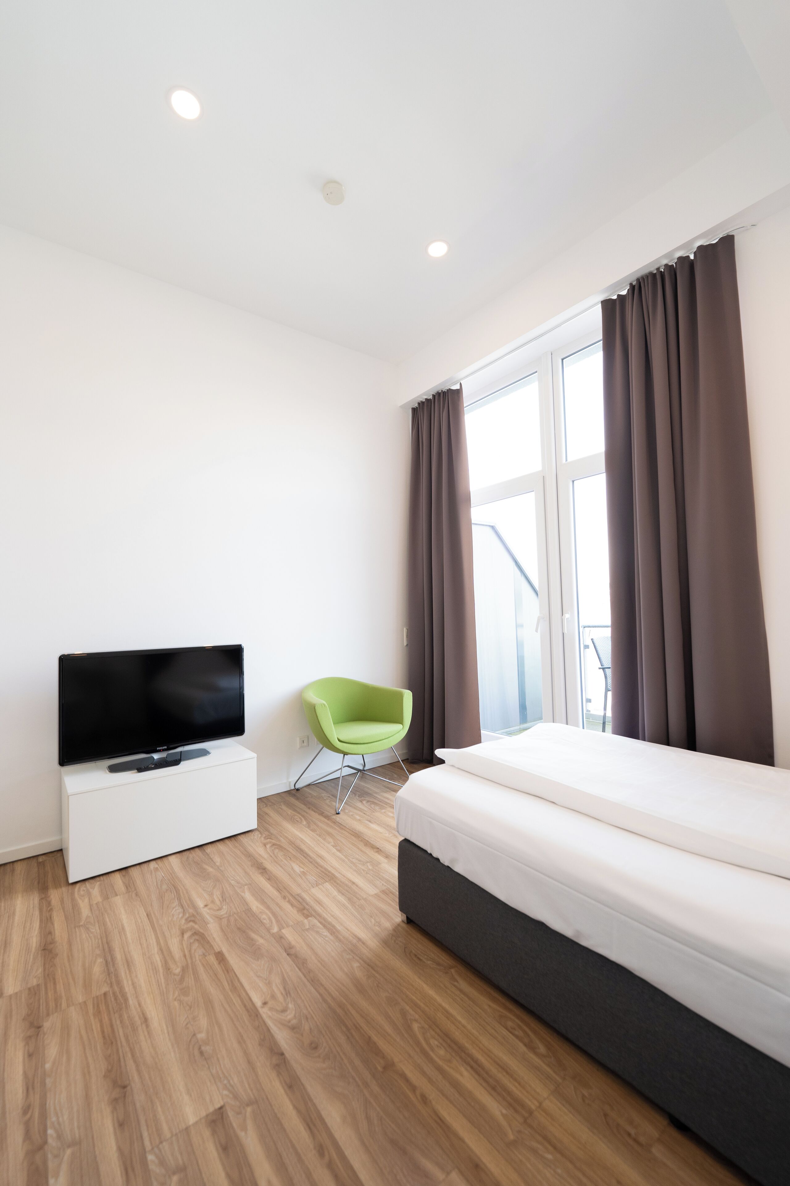 Habitación de hotel moderna con un televisor de pantalla plana sobre un soporte blanco, una silla verde, una cama con cabecera oscura y un ventanal con cortinas.