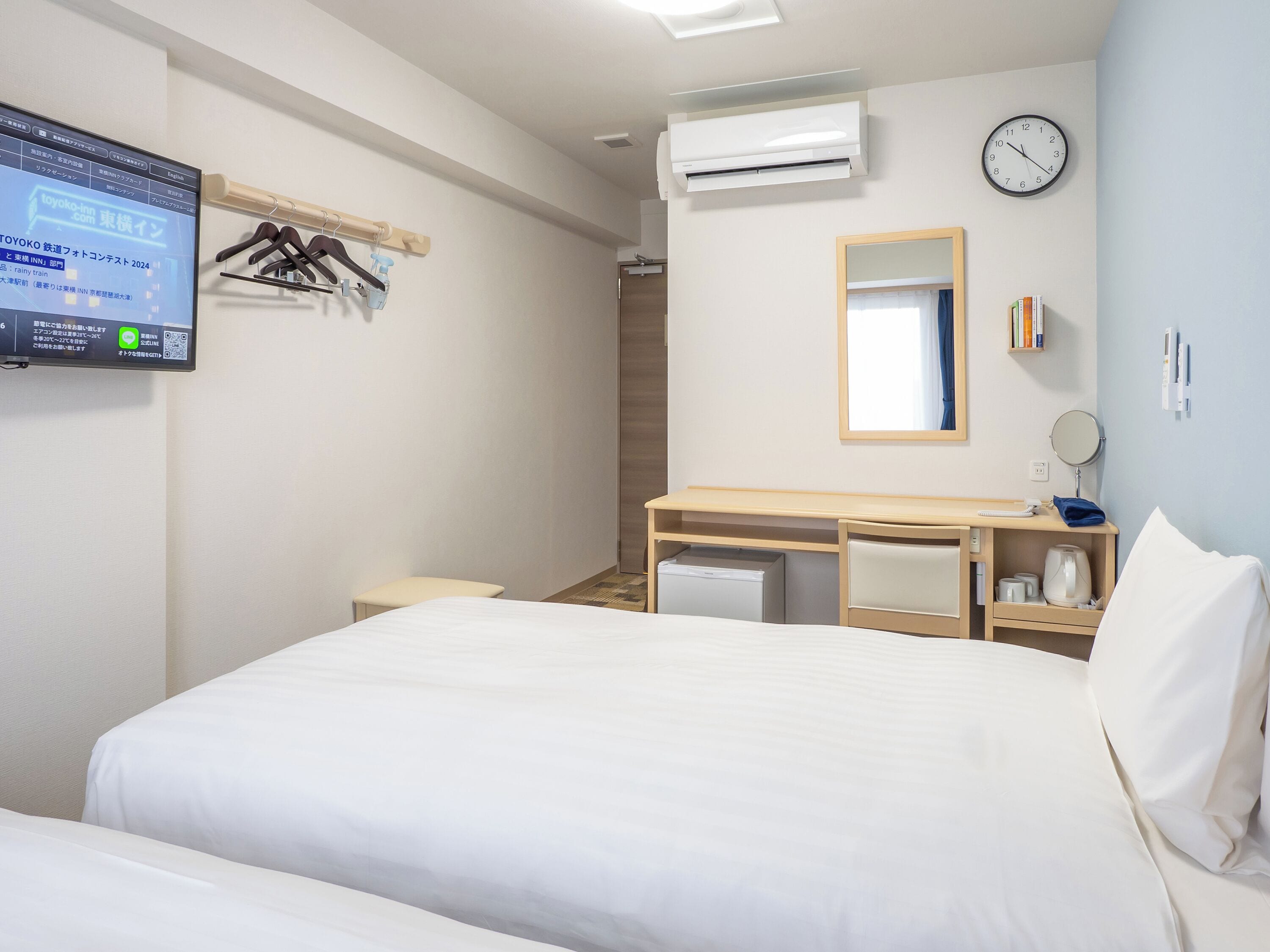 A hotel room with a bed, a desk, a mirror, an air conditioner, and a wall-mounted TV.
