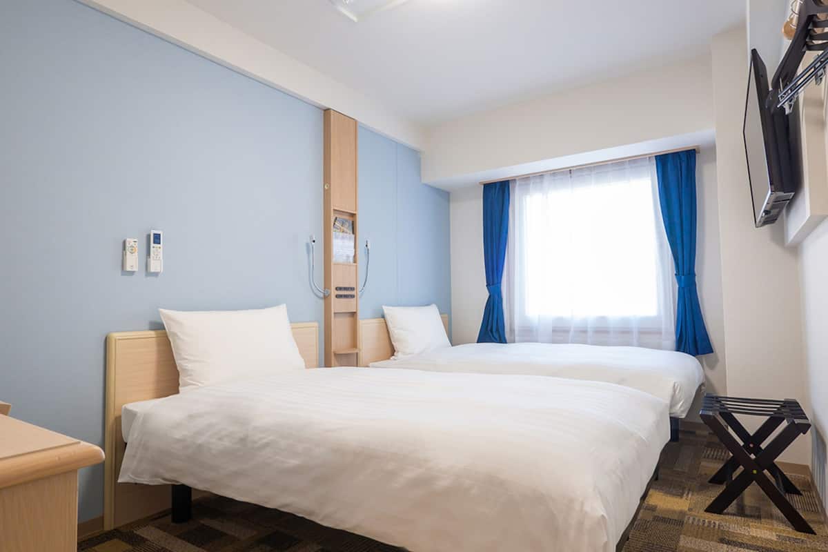 Une chambre d’hôtel avec deux lits, une armoire en bois, une table de chevet, un téléviseur fixé au mur et une fenêtre avec des rideaux bleus.