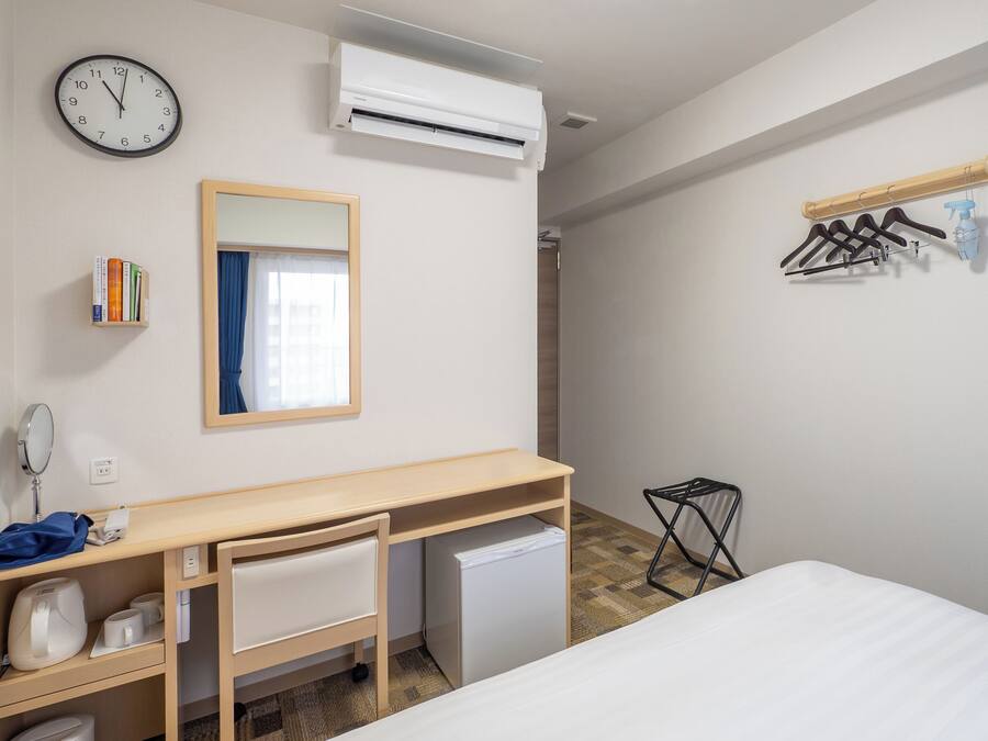 A hotel room with a wooden desk, a small refrigerator, a bed, a wall clock, an air conditioner, a mirror, a window with curtains, a bookshelf, and a coat rack with hangers.