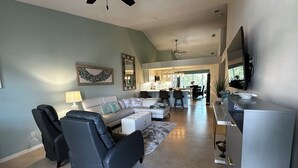 A spacious living room with a sofa, chairs, and a TV. A kitchen with a dining area is visible in the background. - 2 Bed + Den condo in charming Naples with, private pool, WiFi. Relax and unwind (Naples)