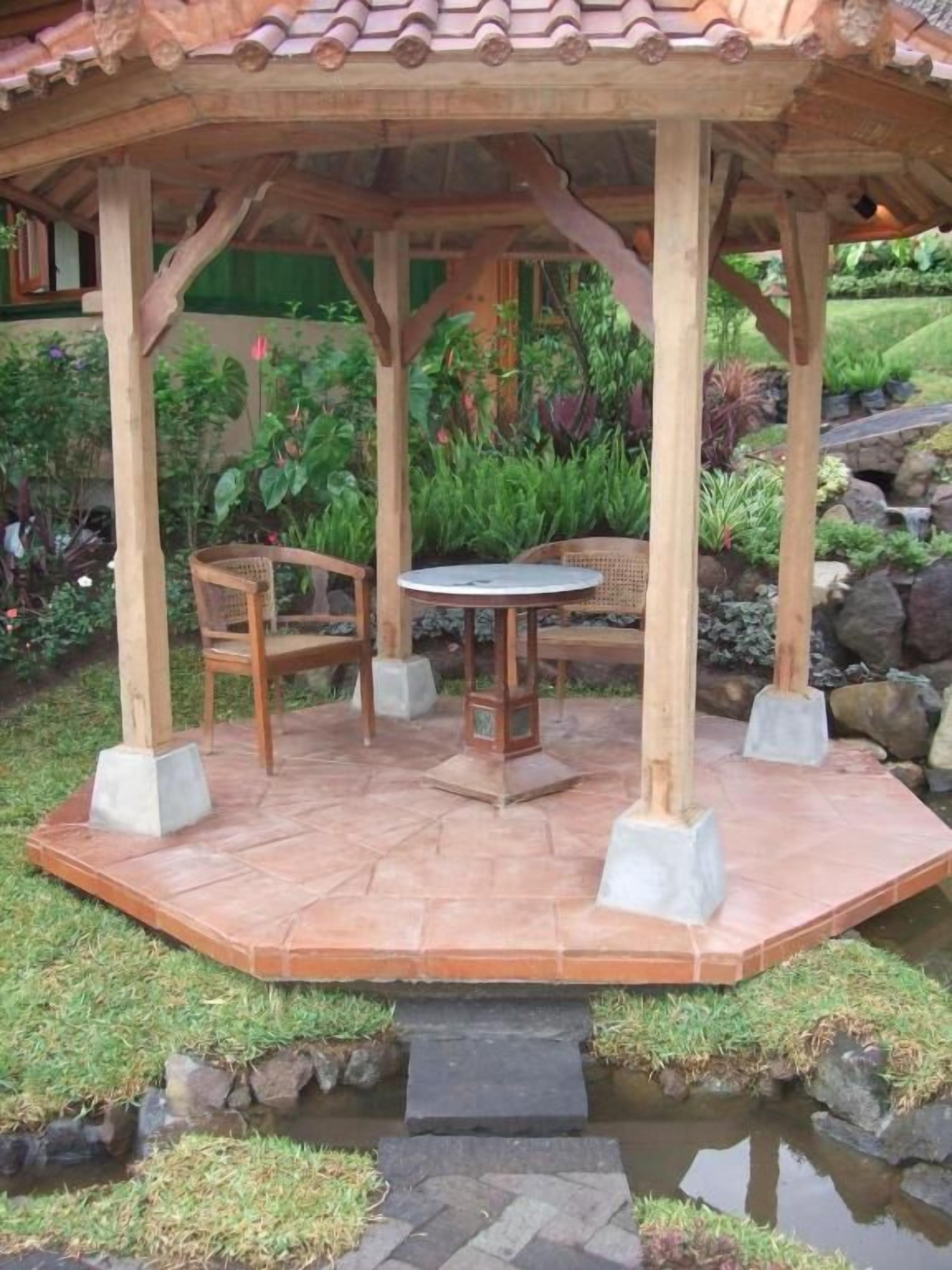 Un quiosco de madera con piso de baldosas, rodeado por un jardín con diversas plantas y un pequeño elemento acuático.