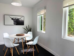 A modern dining area with a round table, white chairs, a framed artwork, and large windows with a view of greenery. - Golden Hour House OB (San Diego)