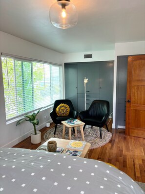 Une chambre à coucher comprenant un lit, une petite table, deux chaises, une plante en pot, une fenêtre avec des stores et une armoire.