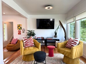A modern living room with two yellow armchairs, a red stool, a black ottoman, a flat-screen TV, and a large window. - Golden Hour House OB (San Diego)