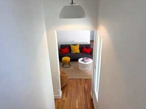 Ein Flur, der in ein Wohnzimmer mit einer Couch, einem runden gelben Hocker und einem weißen Couchtisch mit Büchern führt.