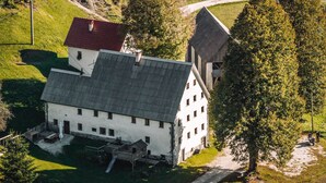 A white building with a sloped roof, a wooden structure to the right, and a paved path leading to it. - Gorenja Trebuša, Domačija Gačnik (Tolmin)