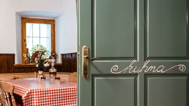 A dining area with a checkered tablecloth, wooden chairs, and a green door with the name "Kuhna" on it. - Gorenja Trebuša, Domačija Gačnik (Tolmin)
