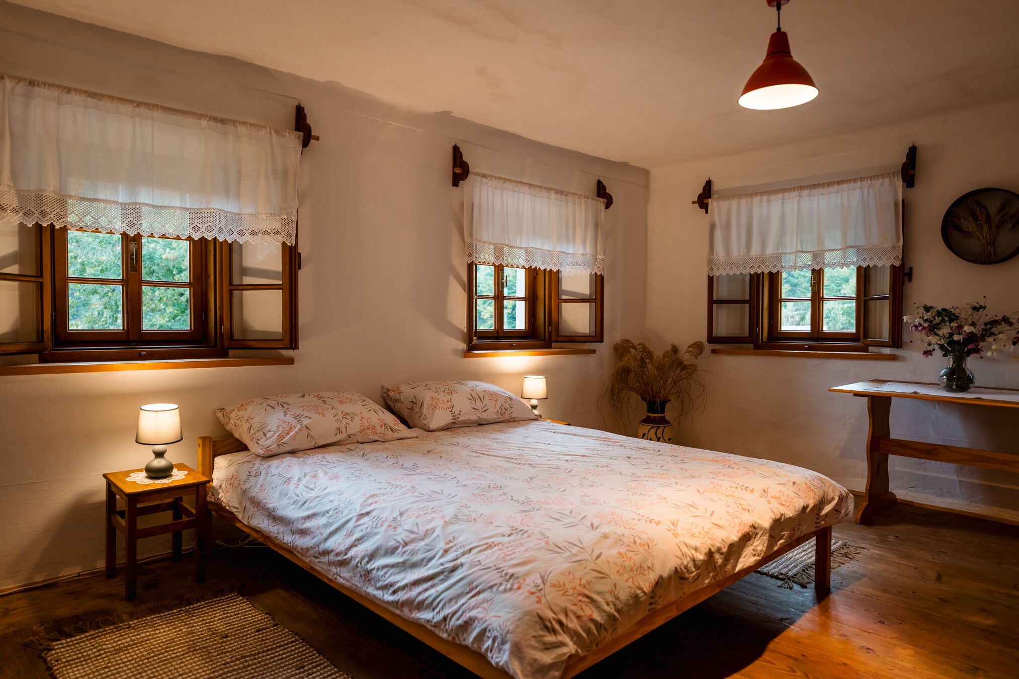 A bedroom with a large bed, two bedside tables with lamps, a wooden desk, and a window with a lace curtain.