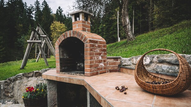 A brick outdoor oven with a chimney, a stone bench, and a woven basket on a tiled patio. - Gorenja Trebuša, Domačija Gačnik (Tolmin)