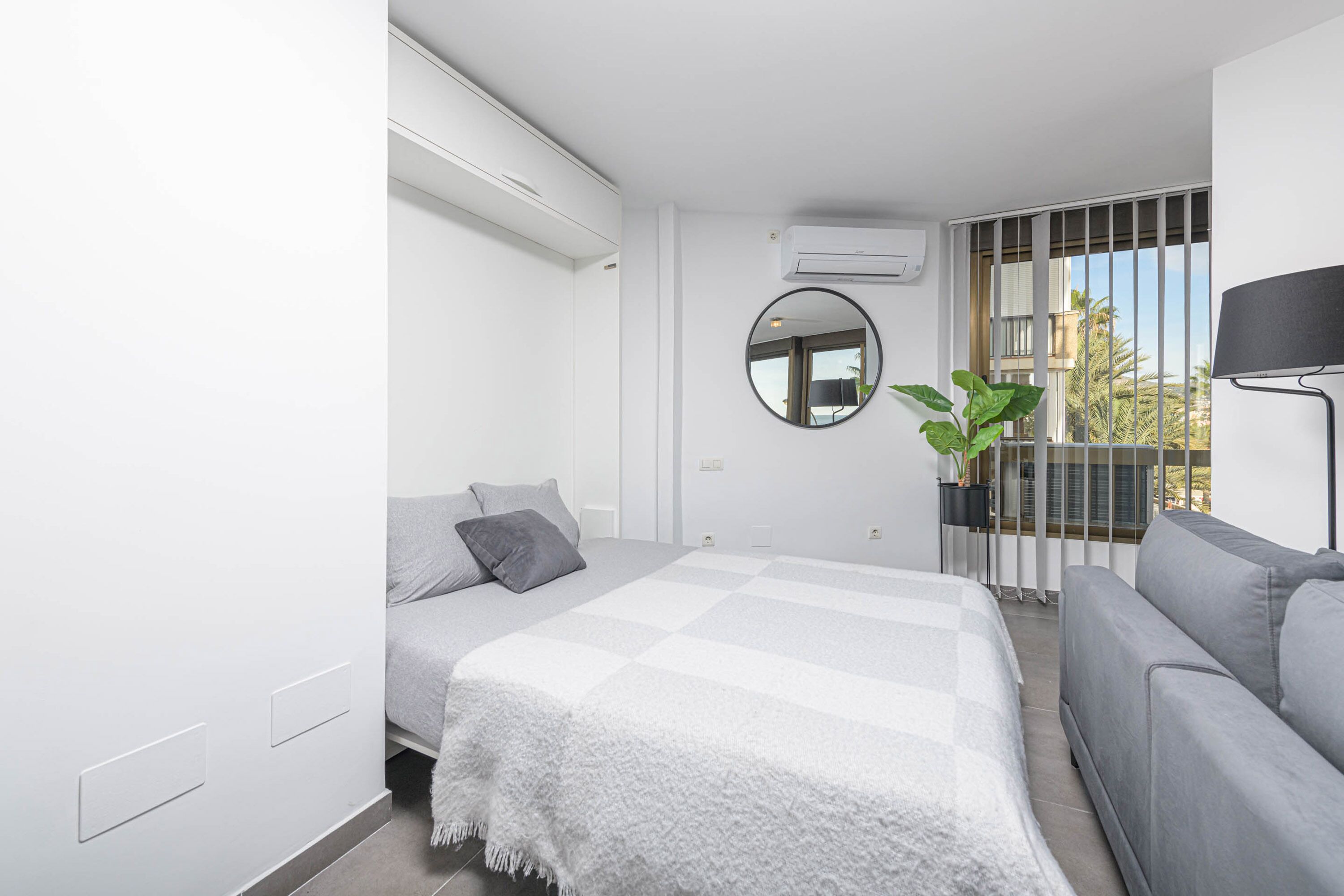 Une chambre à coucher moderne avec un lit, un canapé gris, un miroir rond et une plante en pot.