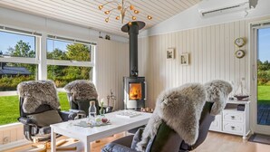 A modern living room with a fireplace, two fur-covered chairs, a dining table, and large windows offering a view of the outdoors. - "Galmand" -  from the sea by Interhome (Hals)
