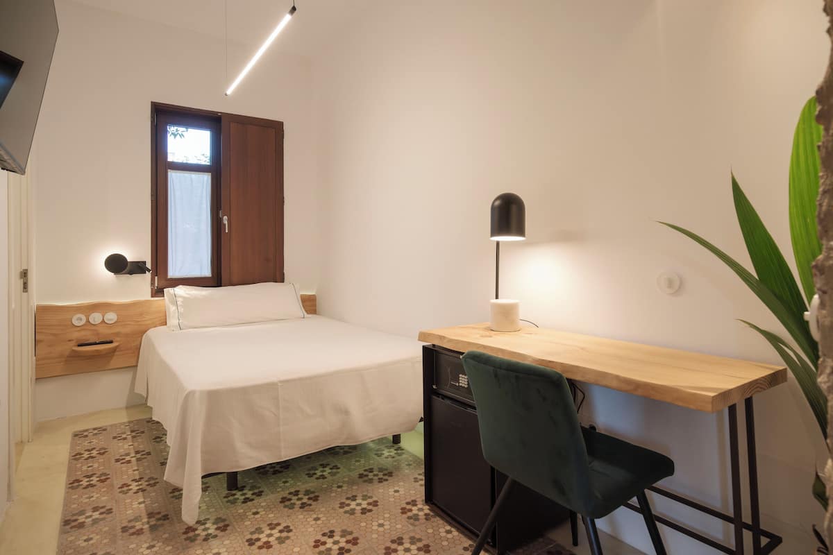 A bedroom with a bed, a desk, a chair, a small refrigerator, and a window with curtains.