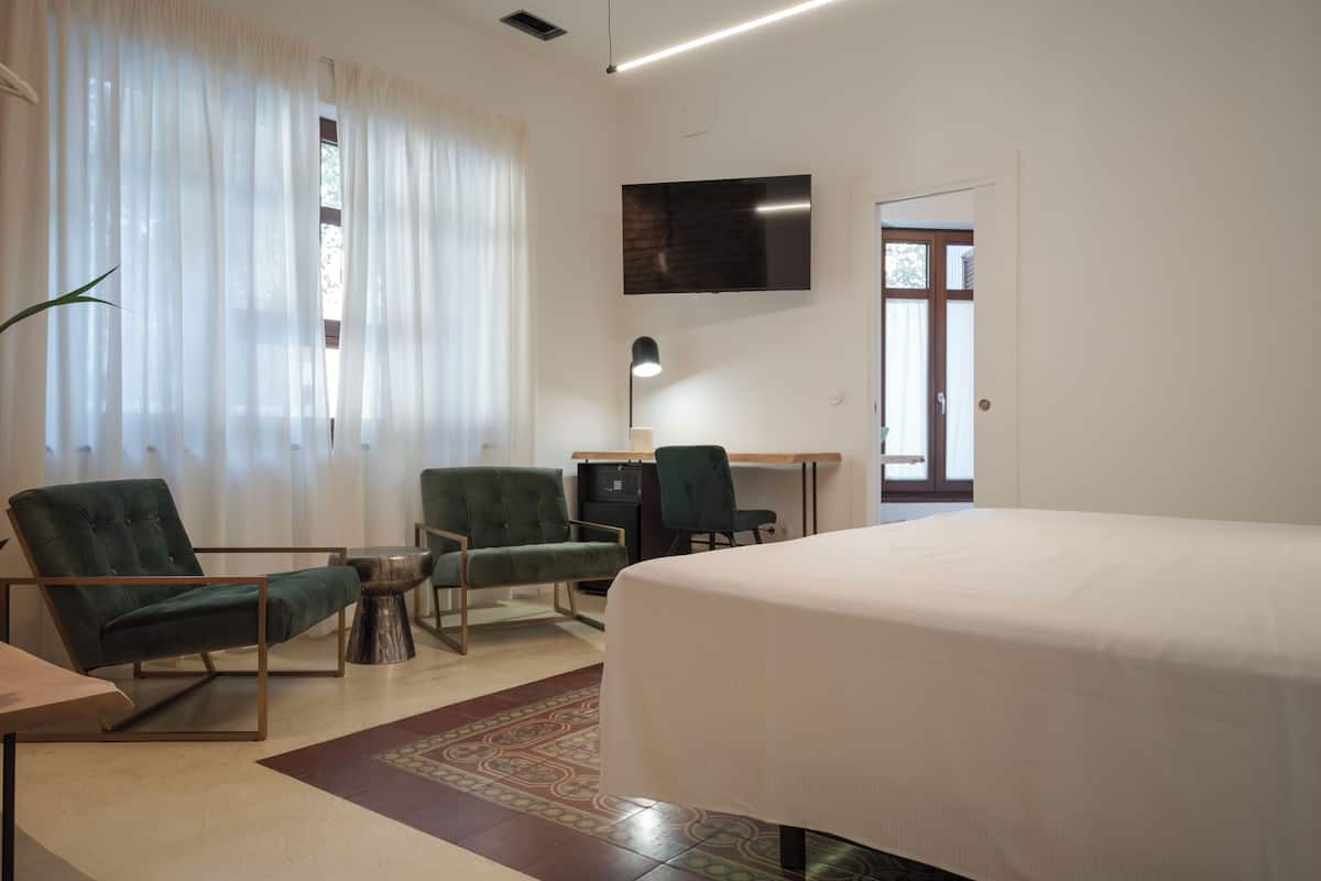 A hotel room with a bed, two green armchairs, a desk with a computer, a TV mounted on the wall, and a large window with sheer curtains.