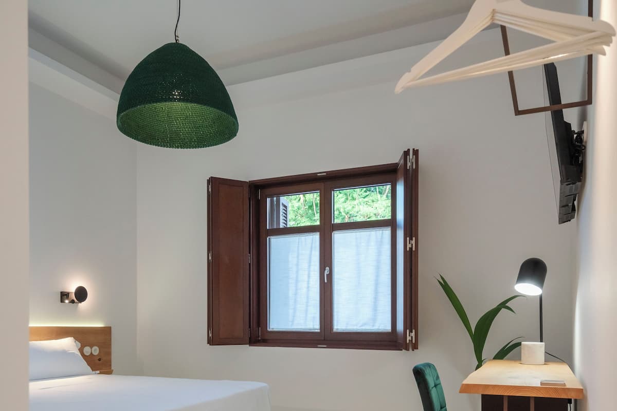 A bedroom with a bed, a desk, a chair, a hanging light, a window, and patterned flooring.