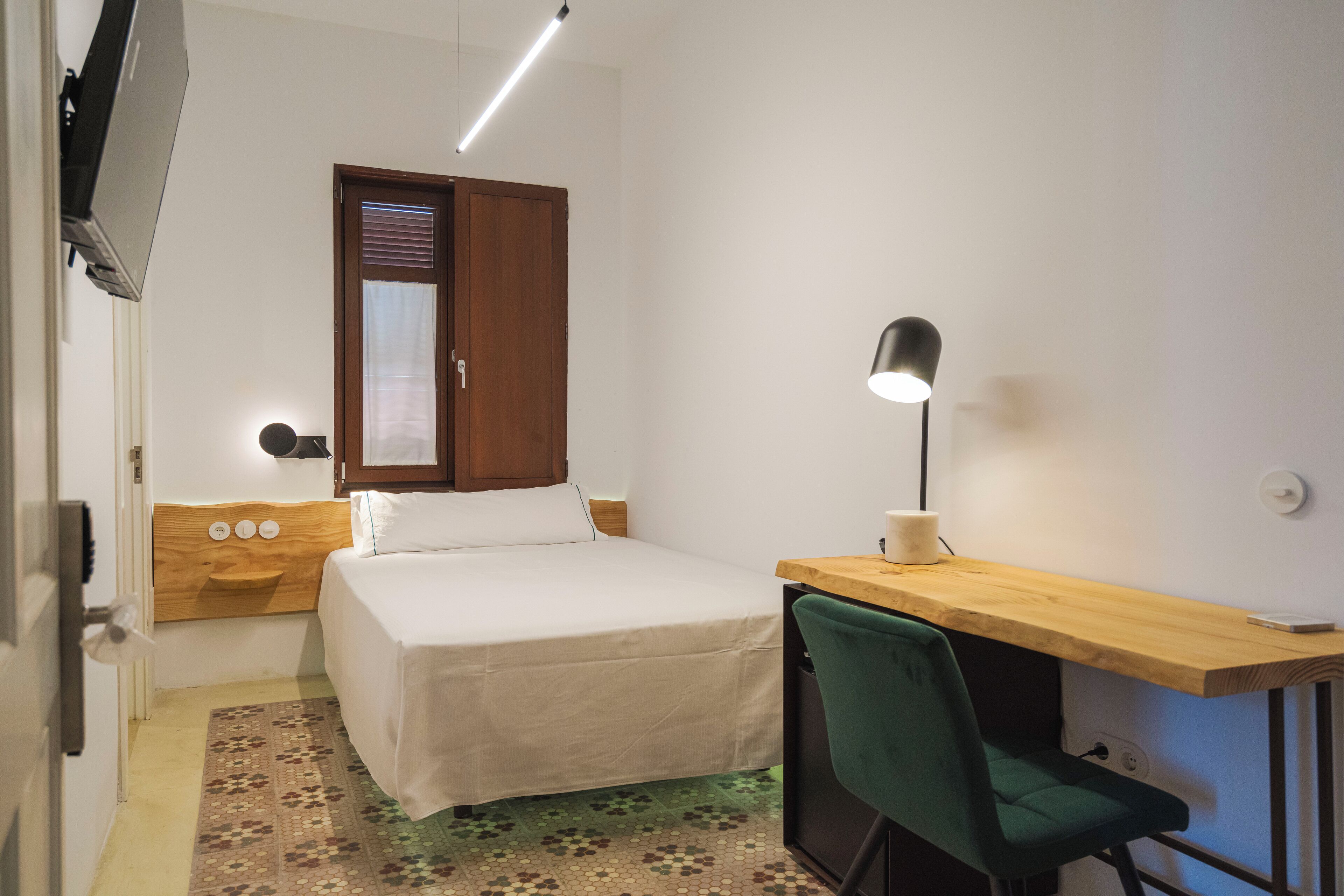 Ein Schlafzimmer mit Bett, Schreibtisch, Stuhl, Lampe, einem Fenster mit Jalousien und einem an der Wand montierten Fernseher.