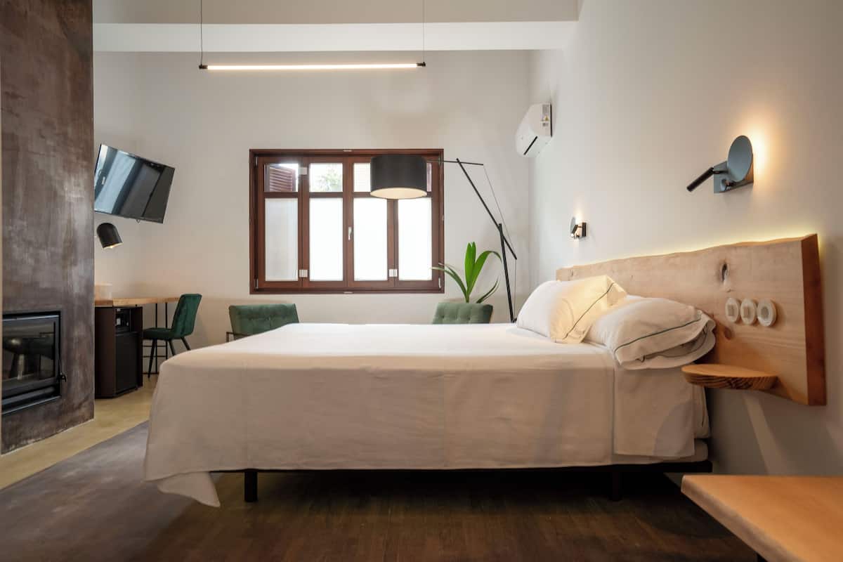 A modern bedroom with a large bed, a TV mounted on the wall, a desk with a chair, and a window with blinds.