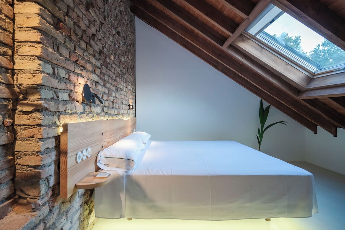 A bedroom with a brick wall, a wooden headboard, a skylight, and a bed with white bedding.