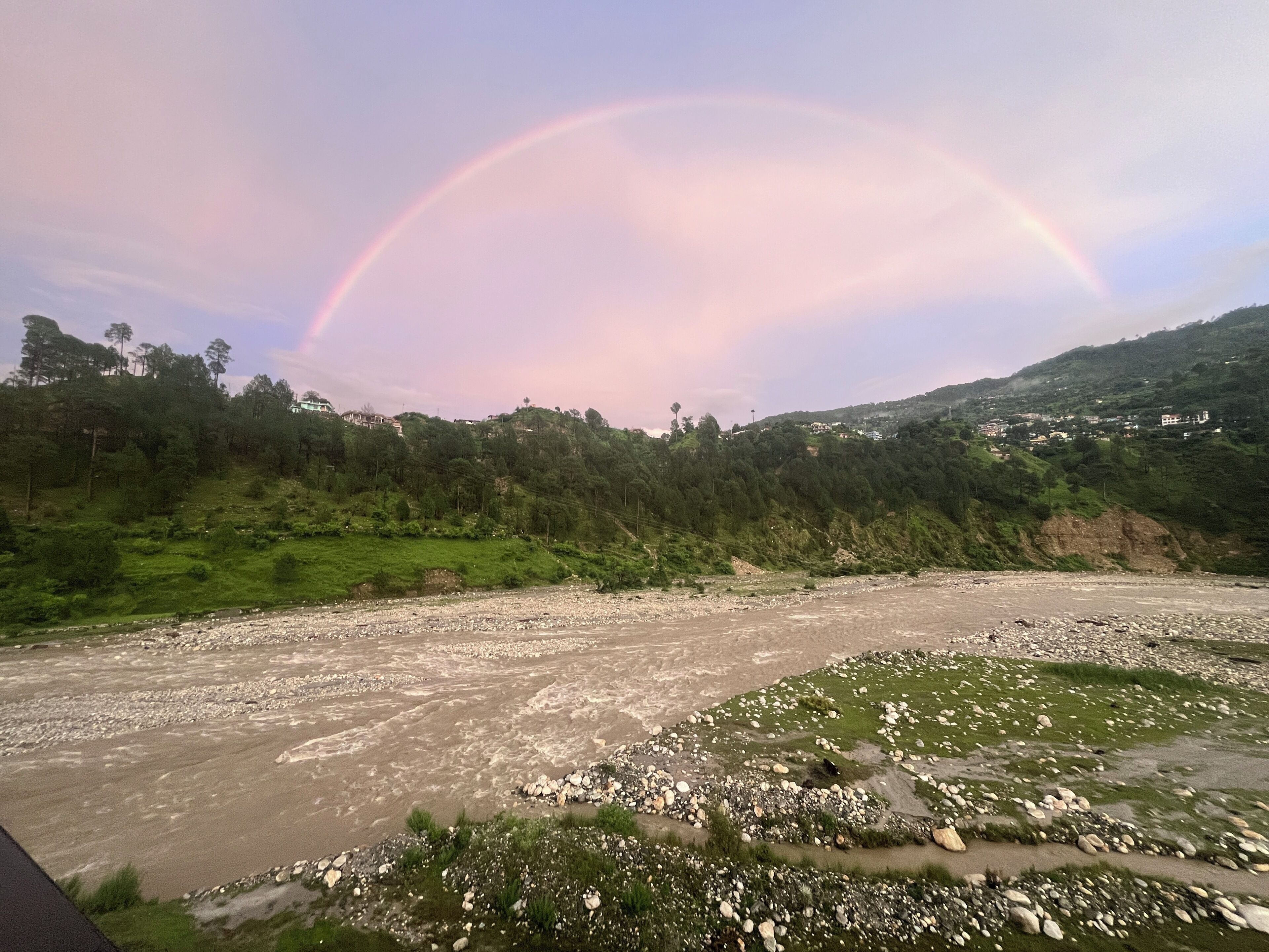 A river with a rainbow, surrounded by green hills and scattered houses.