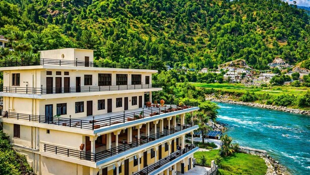 Um hotel de vários andares com varandas a dar para um rio, rodeado por vegetação exuberante e colinas.